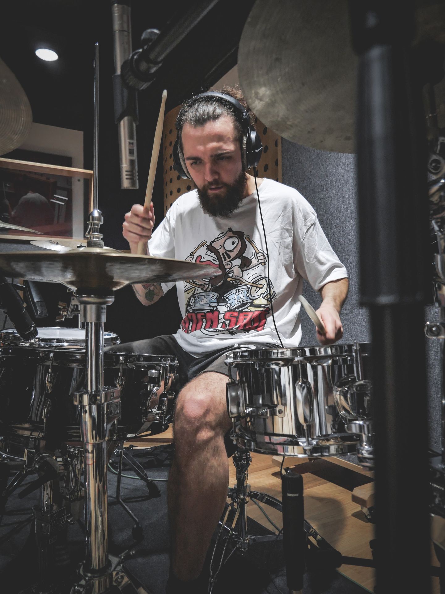 Man playing drums in a music studio wearing headphones and a graphic t-shirt.